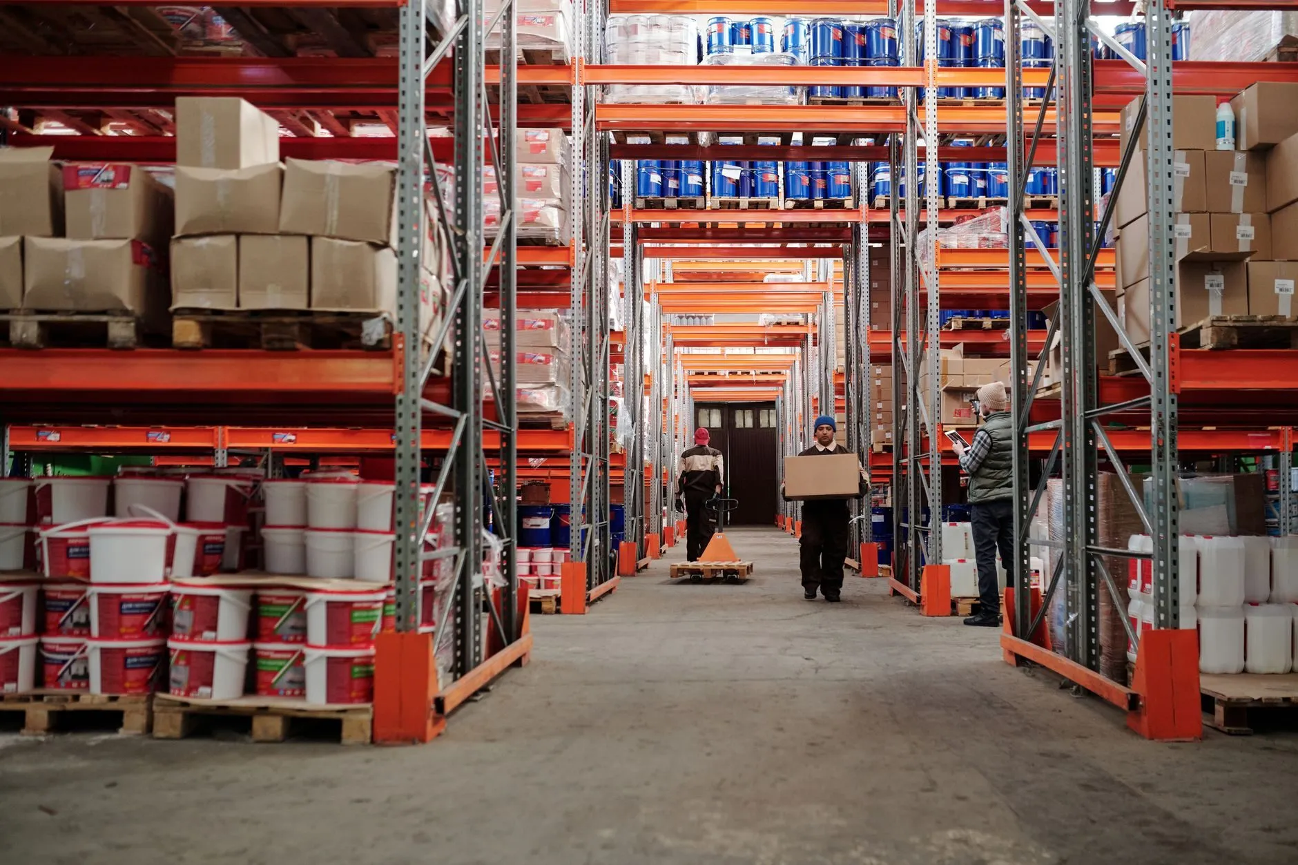 A long warehouse aisle with pallets of stock on orange racking and staff at the far end
