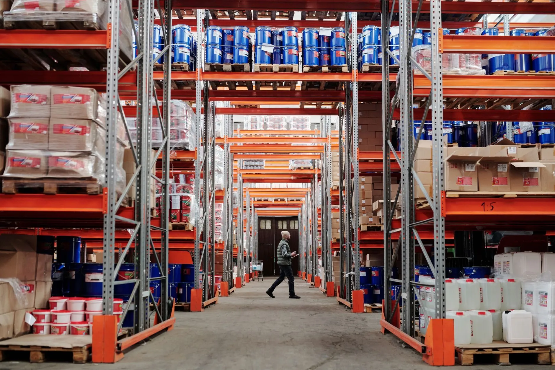 A long warehouse aisle between orange pallet racking, one person walking at the far end