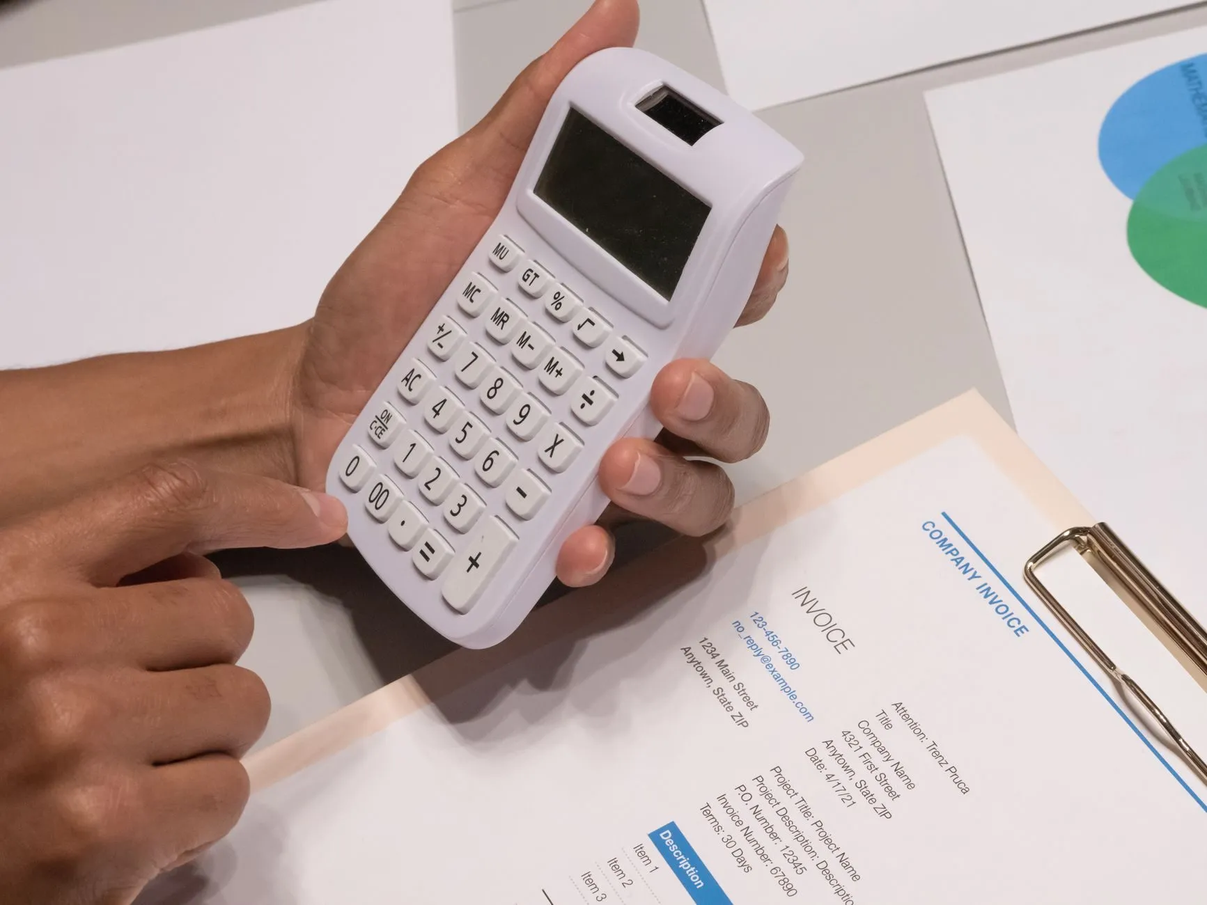 Hands holding a white calculator over a printed company invoice on a clipboard