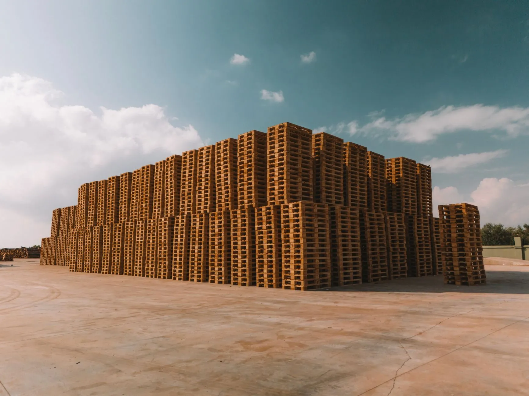 A large block of stacked wooden pallets in an open yard under a cloudy sky